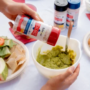 Person sprinkling Tajín seasoning on guacamole in a bowl.